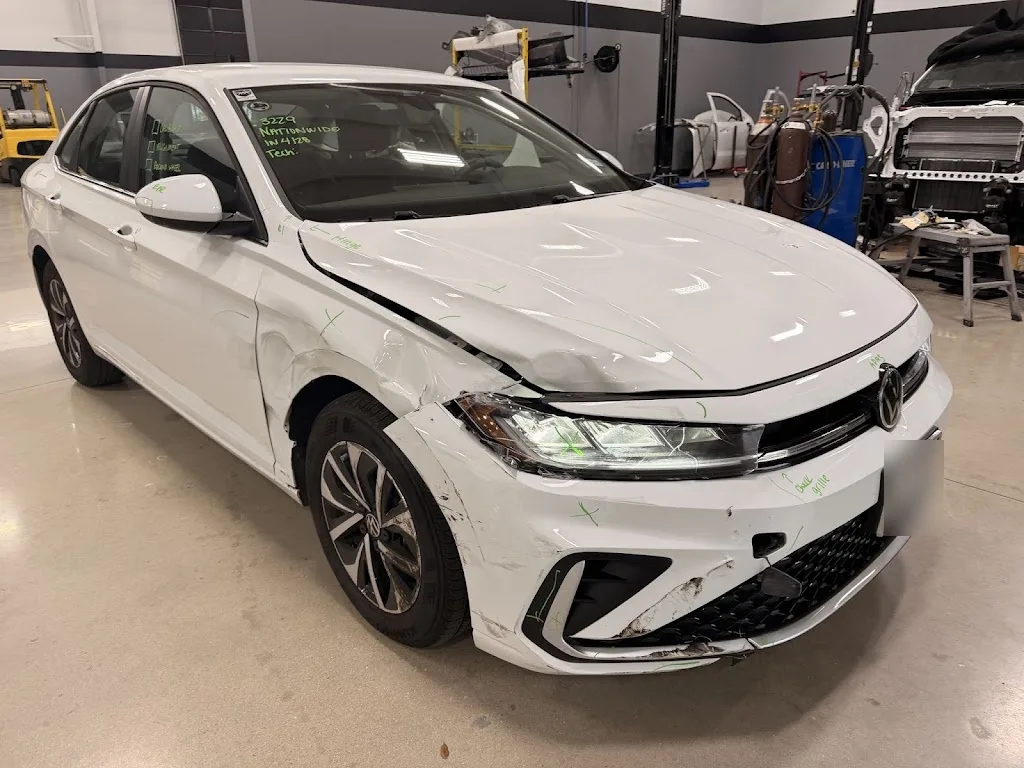White Volkswagen Jetta on the shop floor with front-left collision damage across the fender, bumper cover and headlight, marked up for repair